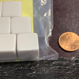May include: A close-up of a package of white plastic dice. The dice are in a clear plastic bag and are arranged in a grid pattern. A penny is visible in the background.