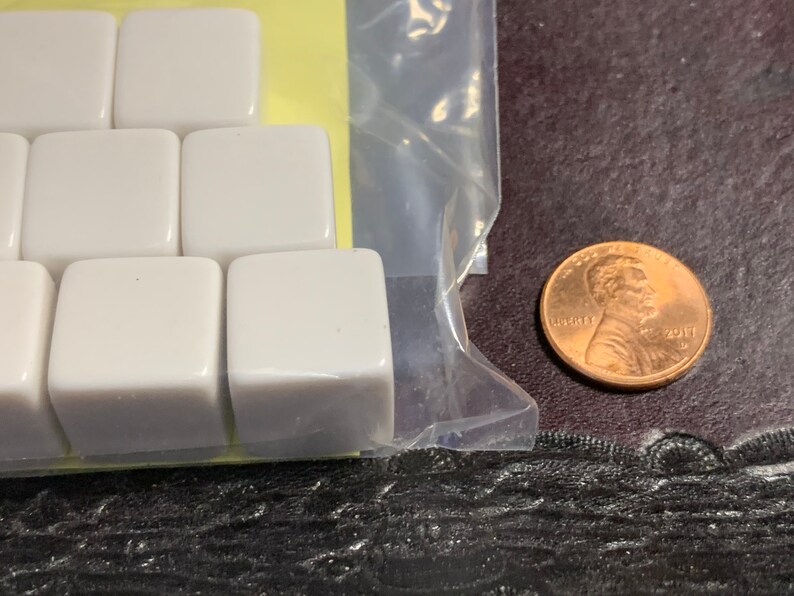 May include: A close-up of a package of white plastic dice. The dice are in a clear plastic bag and are arranged in a grid pattern. A penny is visible in the background.