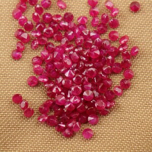 May include: A collection of numerous small, round, faceted rubies with a vibrant deep pinkish-red color. Each gemstone features a brilliant cut with multiple reflective surfaces, arranged in a loose cluster on a textured tan fabric surface.