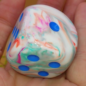 May include: A six-sided die with a smooth, glossy finish featuring a marbled pattern of white, pink, orange, and teal swirls. The die has rounded corners and recessed circular pips in a vibrant blue color across its visible faces.