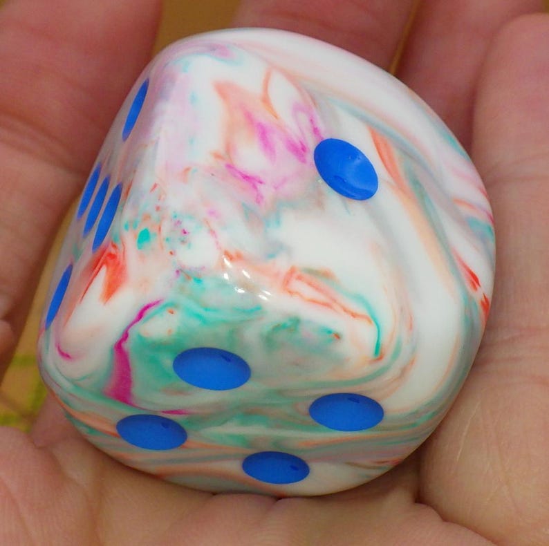 May include: A six-sided die with a smooth, glossy finish featuring a marbled pattern of white, pink, orange, and teal swirls. The die has rounded corners and recessed circular pips in a vibrant blue color across its visible faces.