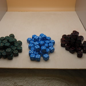May include: Three piles of dice in green, blue, and black. The green dice have gold pips, the blue dice have white pips, and the black dice have red pips. The dice are standard six-sided dice, perfect for tabletop games.