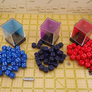May include: An assortment of dice in blue, purple, and red, each with white pips, are displayed. The dice are grouped in piles, with some dice inside clear, angled display boxes. The background features a grid with measurement markings.