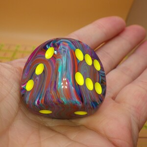May include: A six-sided die featuring a multi-colored marbled pattern with swirls of blue, purple, red, and teal. The die has rounded edges and bright yellow circular pips indicating the numbers, contrasting against the dark, glossy surface.