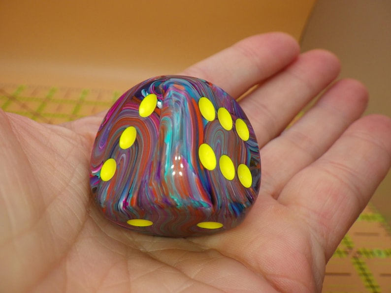 May include: A six-sided die featuring a multi-colored marbled pattern with swirls of blue, purple, red, and teal. The die has rounded edges and bright yellow circular pips indicating the numbers, contrasting against the dark, glossy surface.