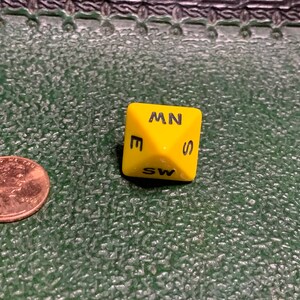 May include: A yellow six-sided die with black letters on each side. The letters are E, SW, S, MN, W, and NW.