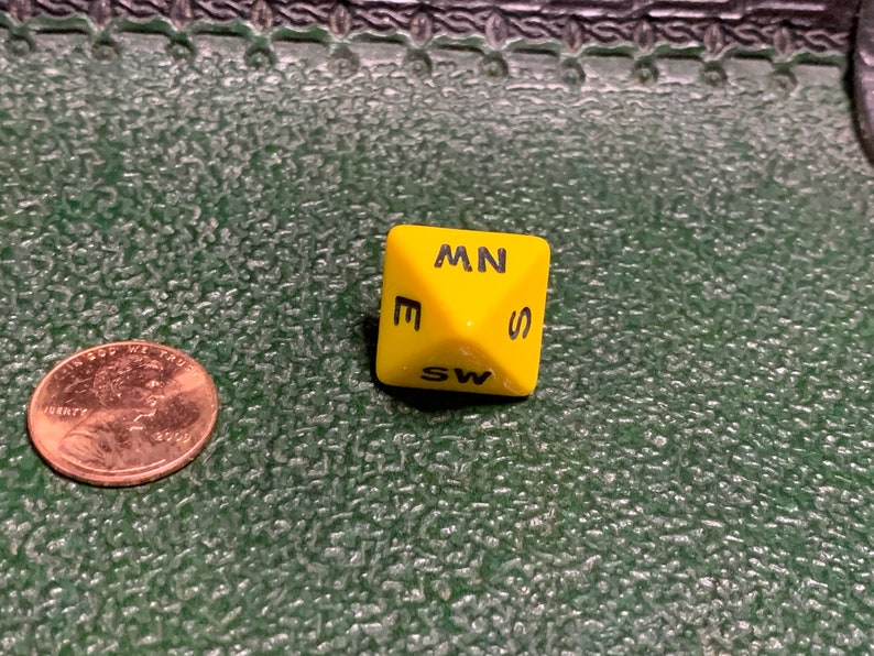 May include: A yellow six-sided die with black letters on each side. The letters are E, SW, S, MN, W, and NW.
