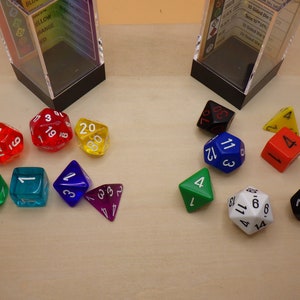 May include: A collection of colorful dice, including red, blue, green, yellow, black, and white. The dice are made of plastic and have different numbers of sides. Some of the dice are in clear plastic containers.
