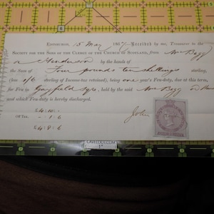 May include: An antique document from 1867, featuring handwritten text and a postage stamp. The document, addressed to Mr. Henderson, details a financial transaction for the Society for the Sons of the Clergy of the Church of Scotland. The document is on a cutting mat.