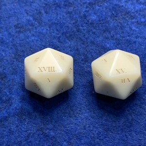May include: Two off-white, twenty-sided dice with Roman numerals. The dice are set against a blue, textured background. Each die has a different Roman numeral visible on its face, indicating its value for tabletop gaming.