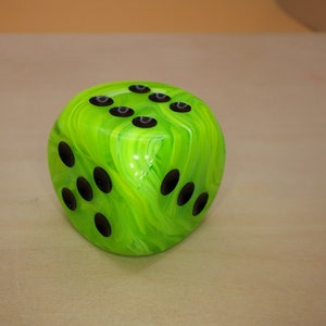 May include: A green and black dice with swirling green and white marbling. The dice has black dots on each side.