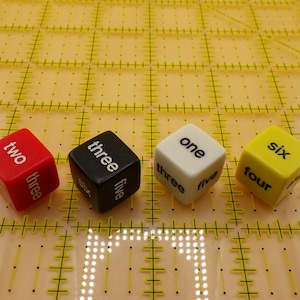 May include: Four colorful dice with numbers and words printed on them. The dice are red, black, white, and yellow. The dice are arranged in a row on a yellow grid.