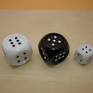 May include: Three dice, one black and two white, with black dots on each face. The dice are on a light brown wooden surface.