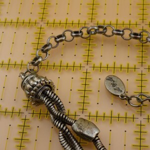 May include: A silver-toned metal necklace featuring a multi-strand snake chain design connected to a textured cylindrical end cap. An oval-shaped metal tag with engraved script text is attached to the chain, which rests on a yellow grid mat.