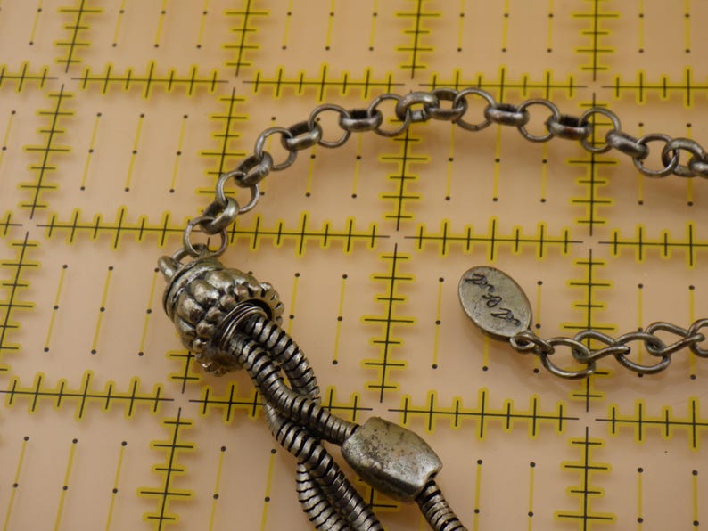 May include: A silver-toned metal necklace featuring a multi-strand snake chain design connected to a textured cylindrical end cap. An oval-shaped metal tag with engraved script text is attached to the chain, which rests on a yellow grid mat.