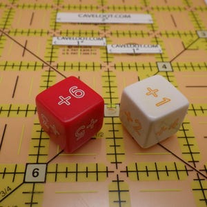 May include: Two dice on a yellow grid. One die is red with white markings, showing +6, +5, and +2. The other die is white with orange markings, showing +1 and +2. The grid has measurements in inches and the text "CAVELOOT.COM".