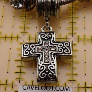 May include: A silver-toned metal cross pendant featuring intricate scrollwork patterns and a central vertical row of five clear rhinestones. The pendant is suspended from a metal bail and is positioned against a yellow grid measuring mat.