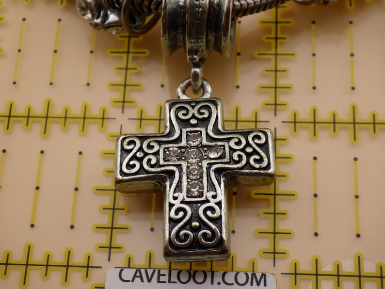May include: A silver-toned metal cross pendant featuring intricate scrollwork patterns and a central vertical row of five clear rhinestones. The pendant is suspended from a metal bail and is positioned against a yellow grid measuring mat.