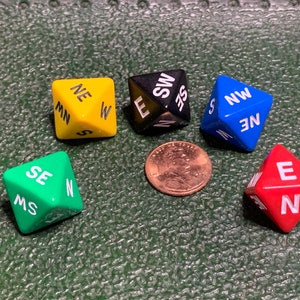 May include: Five different colored dice with the directions North, South, East, West, Northeast, Northwest, Southeast, and Southwest printed on them. The dice are on a green surface.