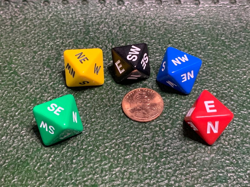 May include: Five different colored dice with the directions North, South, East, West, Northeast, Northwest, Southeast, and Southwest printed on them. The dice are on a green surface.