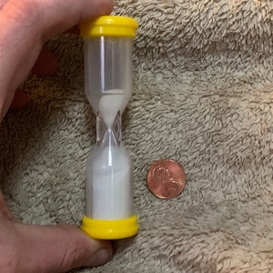 May include: A small, clear hourglass with yellow plastic caps. The hourglass is filled with white sand and is about the size of a US penny.