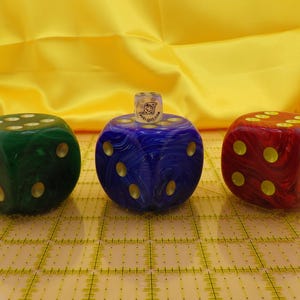 May include: Three large, colorful dice with gold-colored dots. The dice are green, blue, and red, with a small, clear die on top of the blue die. The background is yellow.