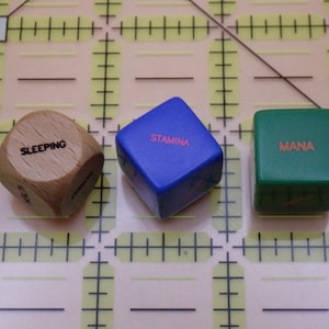 May include: Three dice with different labels. A wooden die with "SLEEPING" in black text, a blue die with "STAMINA" in red text, and a green die with "MANA" in orange text. The dice are on a grid.