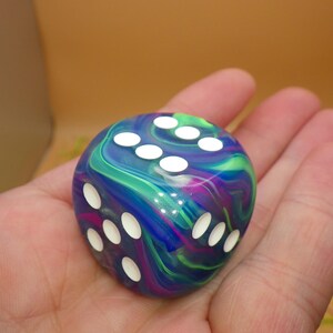 May include: A six-sided die featuring a swirling marbled pattern of deep blue, vibrant purple, and bright green colors. The die has rounded corners and is marked with white circular pips on its faces, showing the numbers three, five, and six.