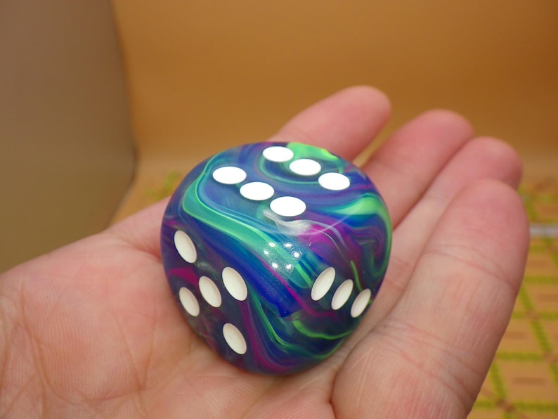 May include: A six-sided die featuring a swirling marbled pattern of deep blue, vibrant purple, and bright green colors. The die has rounded corners and is marked with white circular pips on its faces, showing the numbers three, five, and six.