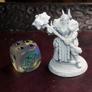 May include: A white painted miniature figurine of a warrior holding a large hammer. The figurine is standing on a wooden base. A clear iridescent dice with black dots and the text "caveloot.com" is in the foreground.