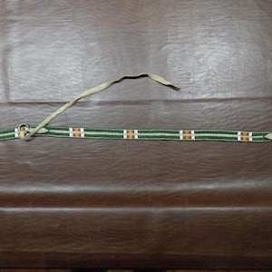 May include: A beaded belt with a green and cream striped pattern. The belt features rectangular brown and white accents. A cream-colored strap is attached to the belt. The belt is lying on a brown surface.