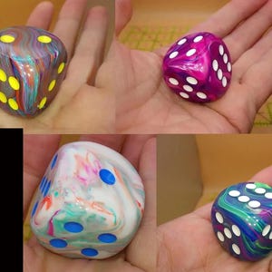 May include: A collection of four six-sided dice featuring marbled, multi-colored resin bodies. One die has a dark base with neon yellow pips, another is magenta and purple with white pips, one is white with pastel swirls and blue pips, and one is blue and green with white pips.