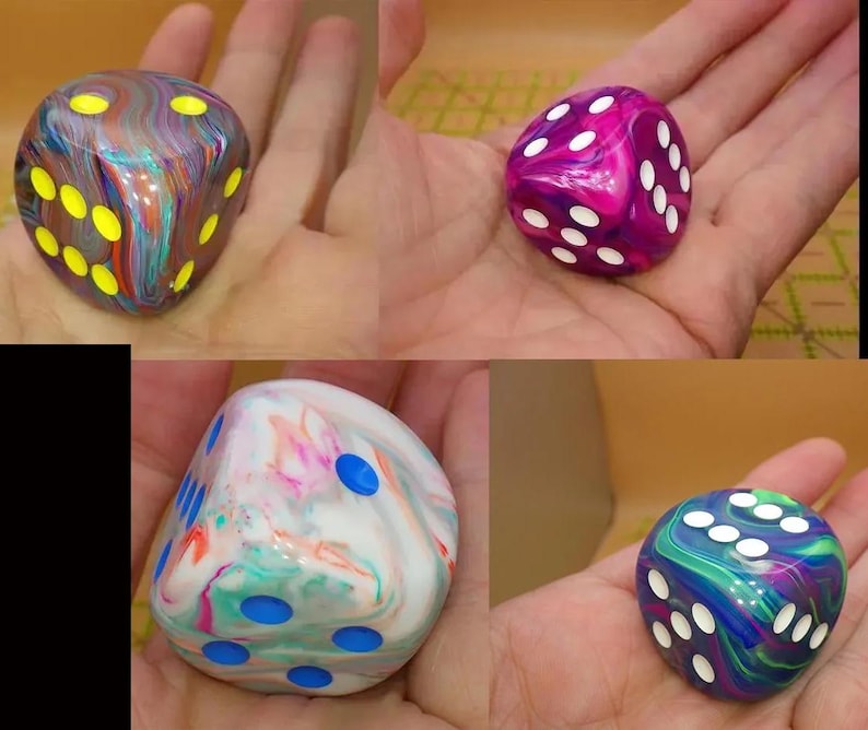 May include: A collection of four six-sided dice featuring marbled, multi-colored resin bodies. One die has a dark base with neon yellow pips, another is magenta and purple with white pips, one is white with pastel swirls and blue pips, and one is blue and green with white pips.