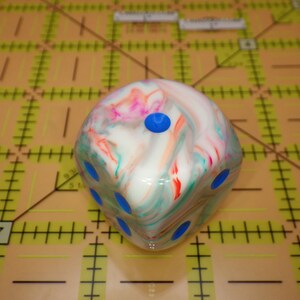 May include: A six-sided die featuring a marbled pattern of white, pink, and teal swirls. The die has bright blue circular pips recessed into each face. It is positioned on a yellow cutting mat with black grid lines and markings in inches.