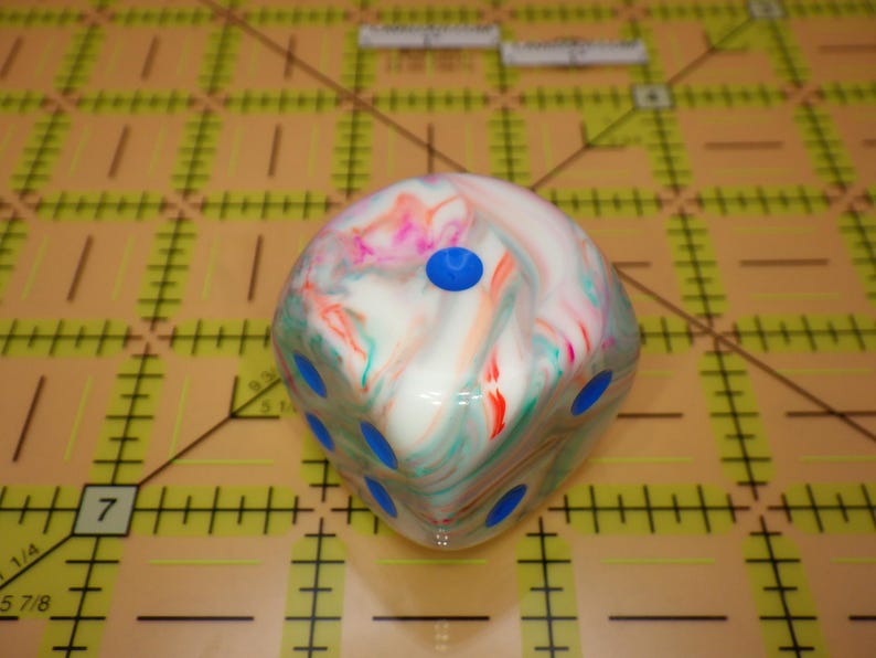 May include: A six-sided die featuring a marbled pattern of white, pink, and teal swirls. The die has bright blue circular pips recessed into each face. It is positioned on a yellow cutting mat with black grid lines and markings in inches.