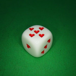 May include: A white dice with red hearts on each side. The dice is on a green surface.
