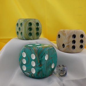 May include: Four decorative dice in various sizes and colors. The largest is teal with white dots, the next is green, and the third is tan. A small, clear die with a logo is also present. The dice are on a white surface with a yellow background.