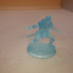 May include: A translucent blue miniature figurine of a person in a fighting pose. The figurine is standing on a round base.