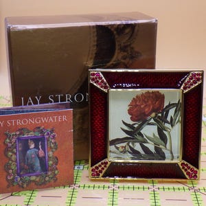 May include: A decorative red and gold picture frame with a floral print, featuring a red flower and green leaves. The frame is embellished with red gemstones. A small booklet and a gold box with the text "JAY STRONGWATER" are also in the image.