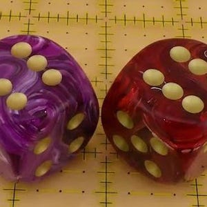 May include: Two six-sided dice, one purple and one red, both featuring a swirling marbled pattern and rounded corners. Each die has cream-colored circular pips recessed into the surface, with the top faces showing a total of six pips each.