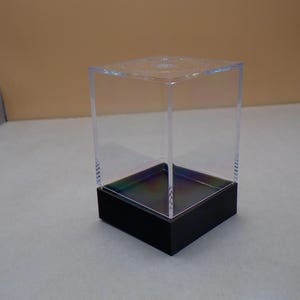 May include: A clear acrylic cube display case with a black base. The case is empty and ready to display a collectible item.