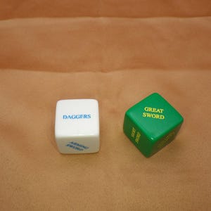May include: Two dice for a tabletop game. One die is white with "DAGGERS" and "ARMING SWORD" in blue text. The other die is green with "GREAT SWORD" in yellow text. The dice are on a tan surface.