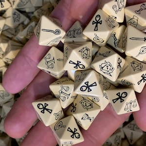 May include: A hand holding a pile of beige triangular dice with black symbols. The symbols include an ankh, a scale, a sword, a chalice, and a rose.