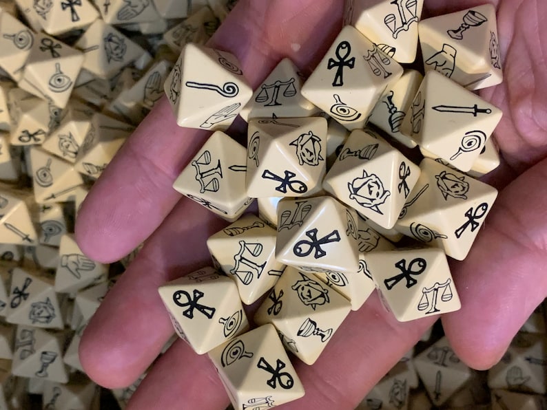 May include: A hand holding a pile of beige triangular dice with black symbols. The symbols include an ankh, a scale, a sword, a chalice, and a rose.