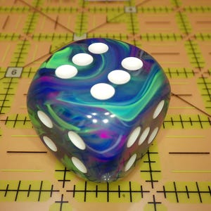 May include: A six-sided die featuring a swirling, marbled pattern of blue, green, and purple colors. The die has rounded corners and white circular pips indicating the numbers. It is positioned on a tan cutting mat with black grid lines.