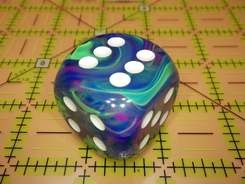 May include: A six-sided die featuring a swirling, marbled pattern of blue, green, and purple colors. The die has rounded corners and white circular pips indicating the numbers. It is positioned on a tan cutting mat with black grid lines.