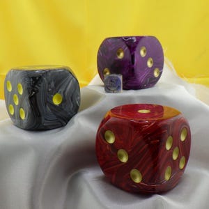 May include: Three large, decorative dice in black, purple, and red with gold-colored dots. A small die is also present. The dice are on a white cloth with a yellow background. The dice are likely for games or decor.