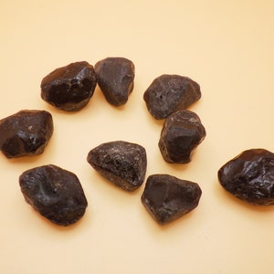 May include: A collection of ten black obsidian stones, each with a unique shape and texture. The stones are arranged in a circle on a light brown surface.