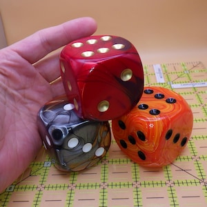 May include: Three large, colorful dice. One is red with gold dots, another is orange with black dots, and the third is black and gray with white dots. The dice are on a grid background.