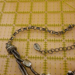May include: A silver-toned multi-strand necklace featuring flexible snake chains, a decorative ribbed bead, and a small oval metal tag with engraved text. The necklace includes a lobster claw clasp and an adjustable chain link extender.